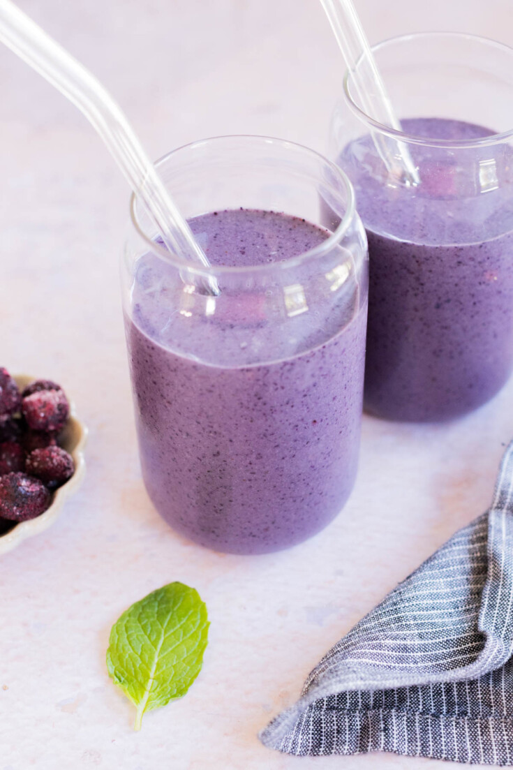 Two clear glasses of blueberry smoothie with glass straws, next to a bowl of frozen berries, a mint leaf, and a striped cloth on a light surface.