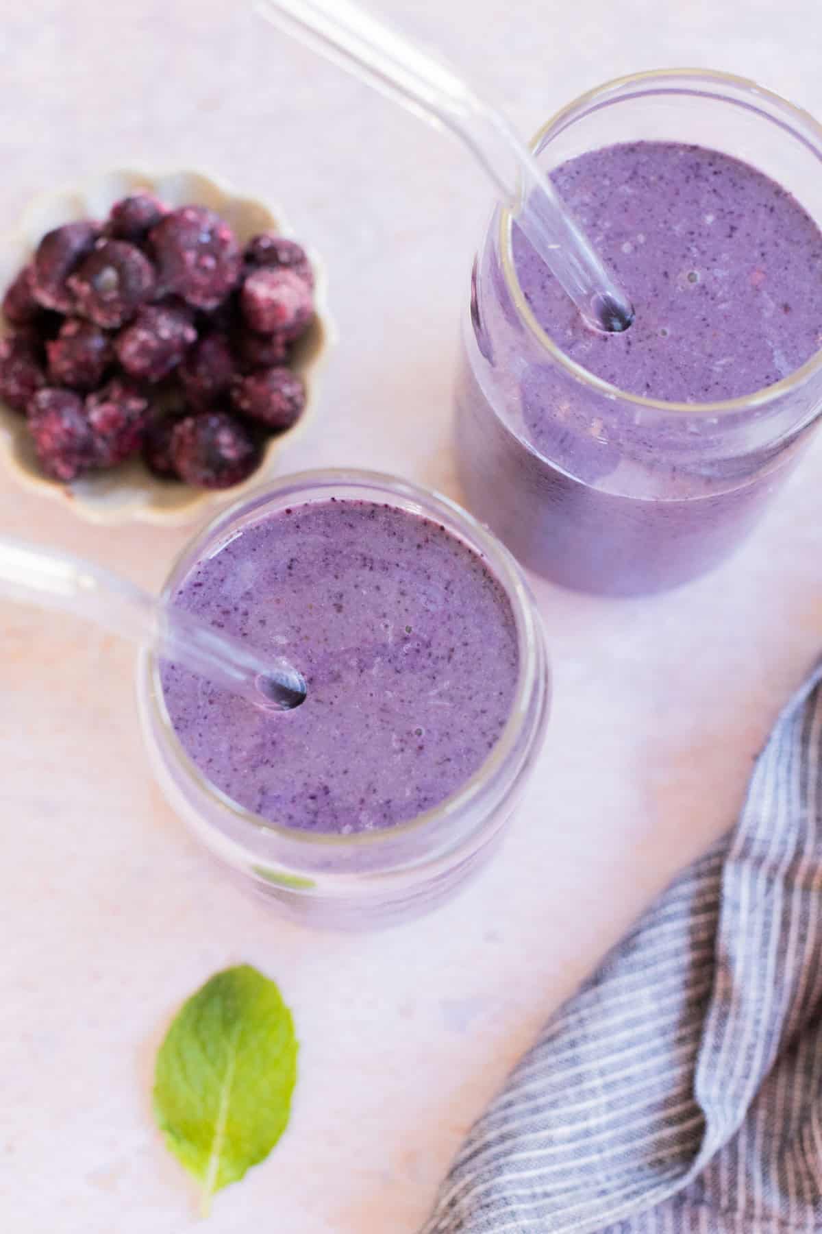 Two blueberry smoothies in glass cups with straws, placed on a light surface. A small bowl of berries and a mint leaf are in the background, with a striped cloth nearby.