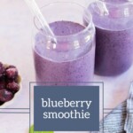 Two glasses of creamy blueberry smoothie with straws rest on a light surface, accompanied by a small bowl of frozen blueberries and a striped cloth. The words "Blueberry Smoothie" elegantly overlay the scene.