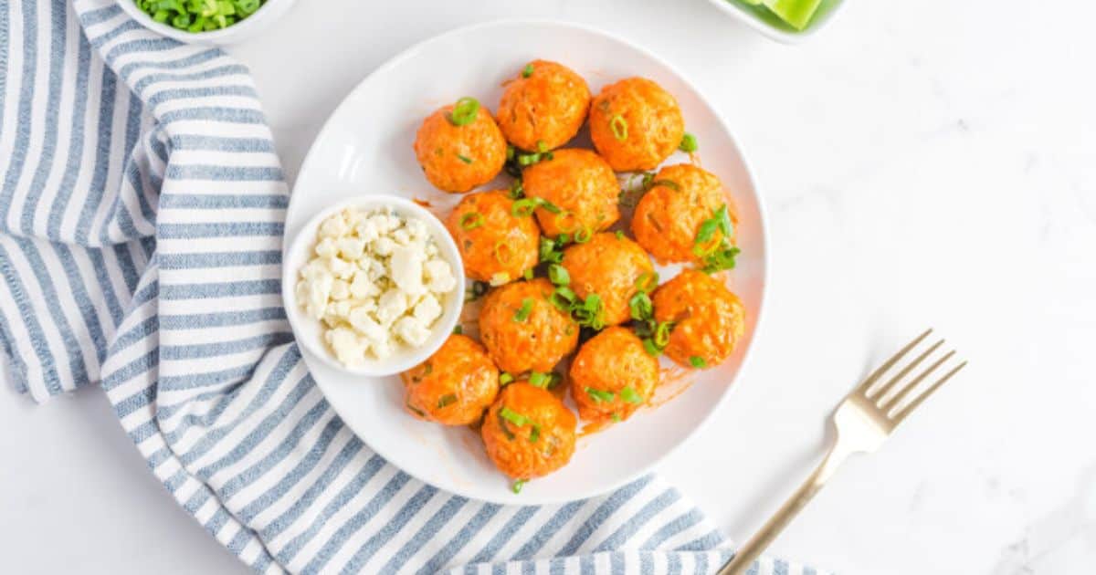 A plate of orange-colored meatballs garnished with green onions, served with a small bowl of crumbled cheese, on a striped cloth next to a fork.