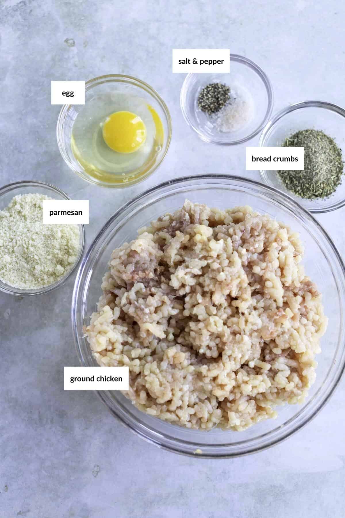 Ingredients for making meatballs on a surface: a bowl of ground chicken, bowls with egg, bread crumbs, grated parmesan, and seasonings labeled salt and pepper.