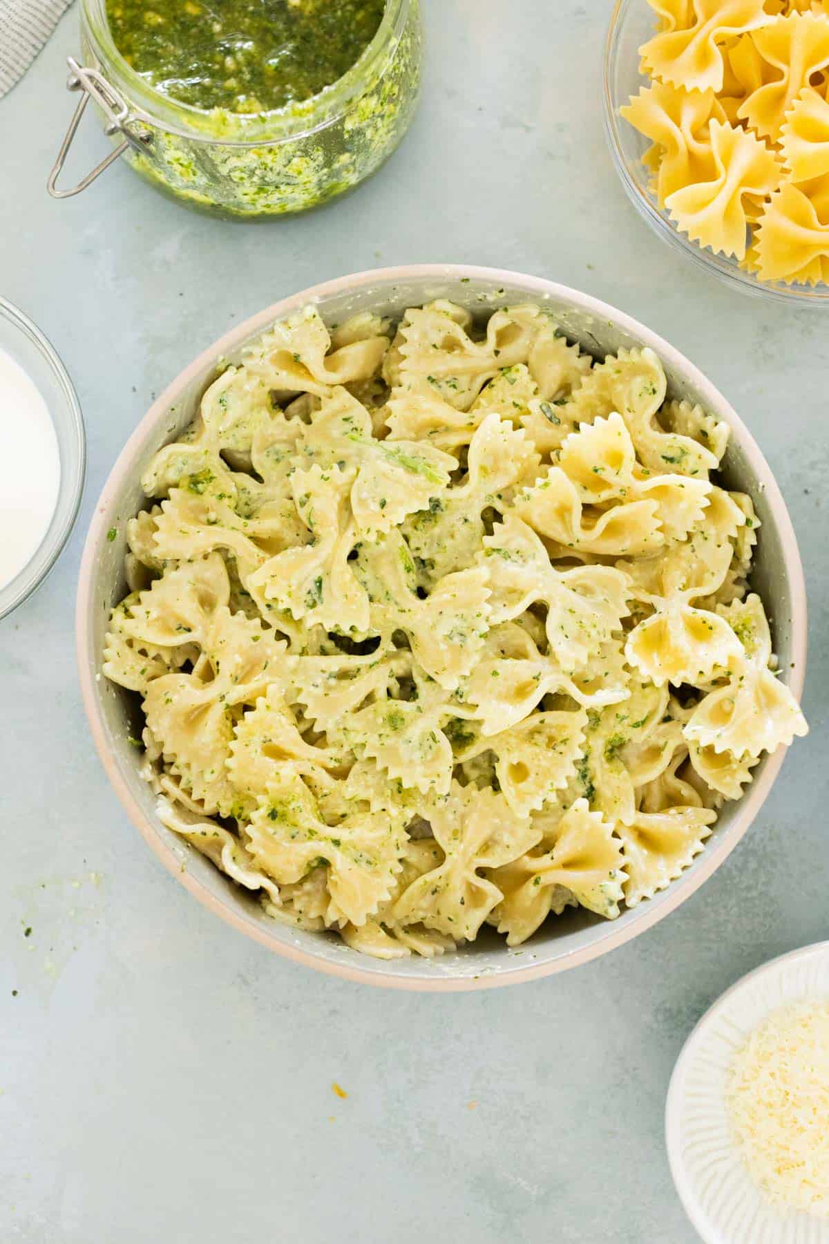 Bowl of bowtie pasta mixed with creamy pesto, surrounded by jars of the pesto sauce and uncooked pasta on a light surface.