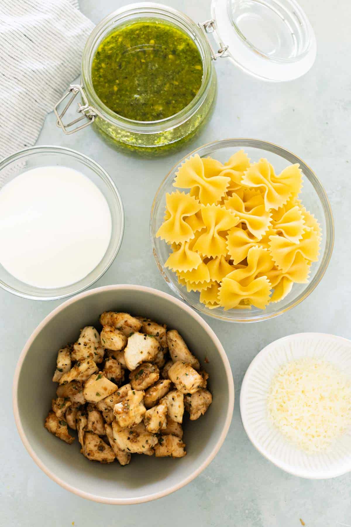 Bowls of pesto sauce, uncooked bowtie pasta, grilled chicken pieces, cream, and grated cheese arranged on a light surface.