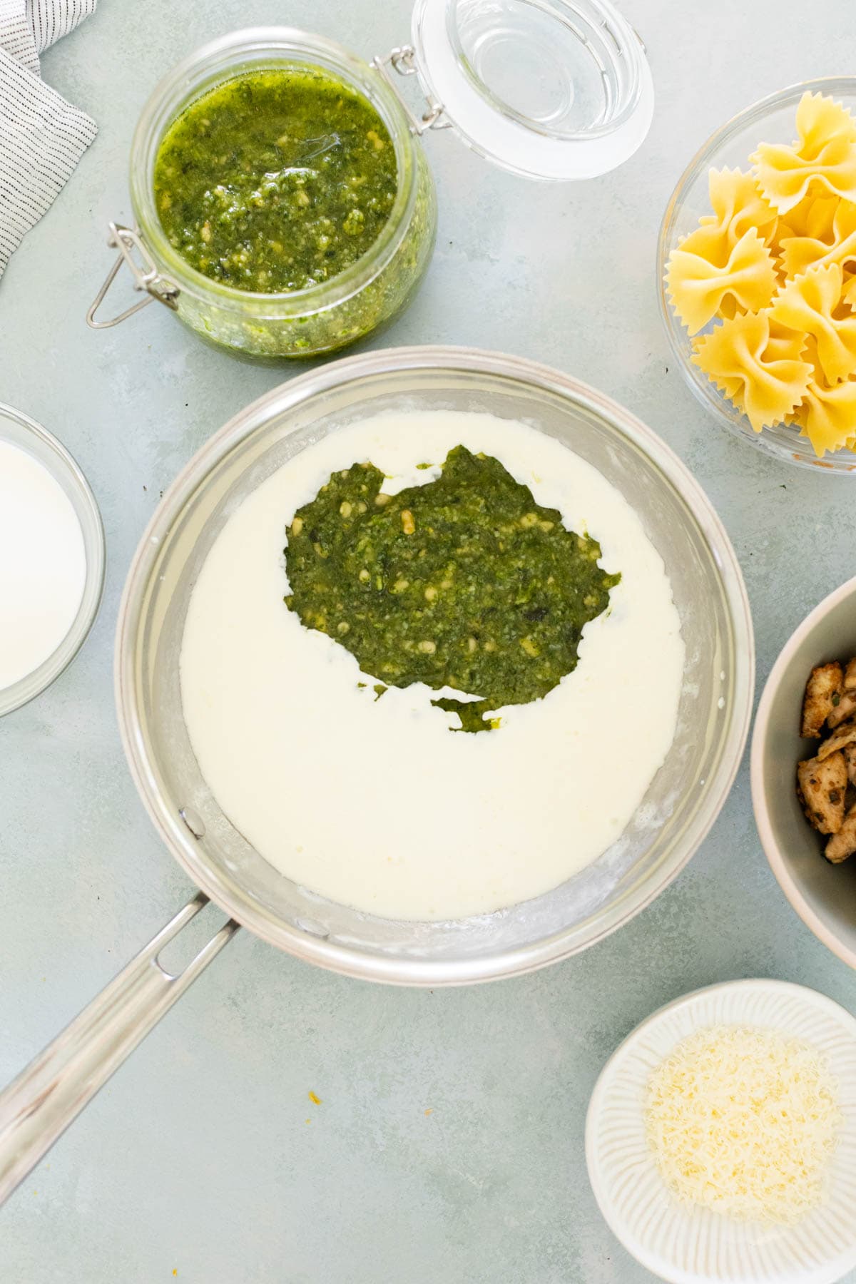 A saucepan with cream and pesto sauce, surrounded by uncooked pasta, cooked chicken, a jar of pesto, and grated cheese on a light surface.