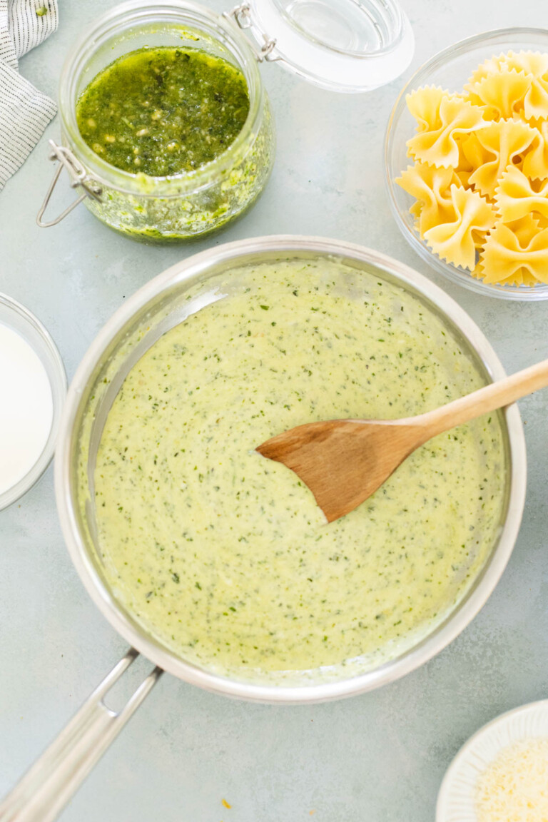 A saucepan with creamy green pesto sauce being stirred with a wooden spoon. Nearby are a jar of pesto and a bowl of uncooked farfalle pasta.