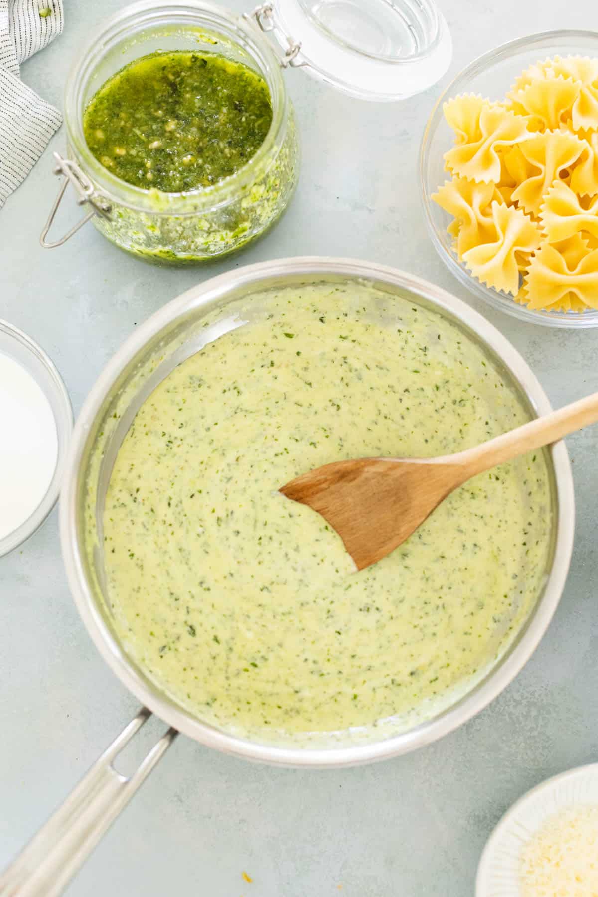 A saucepan with creamy green pesto sauce being stirred with a wooden spoon. Nearby are a jar of pesto and a bowl of uncooked farfalle pasta.