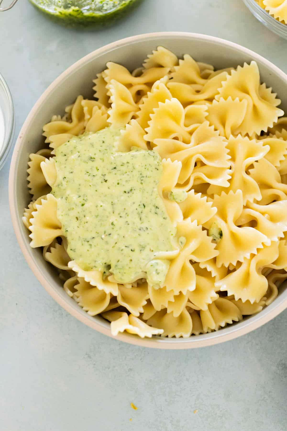 A bowl of bowtie pasta partially covered with green pesto sauce on a light surface.