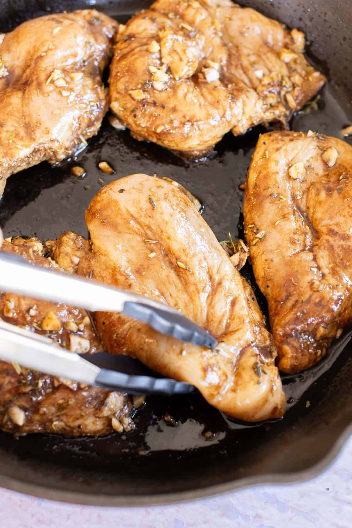 Four pieces of balsamic chicken cooking in a skillet, with metal tongs flipping one piece.