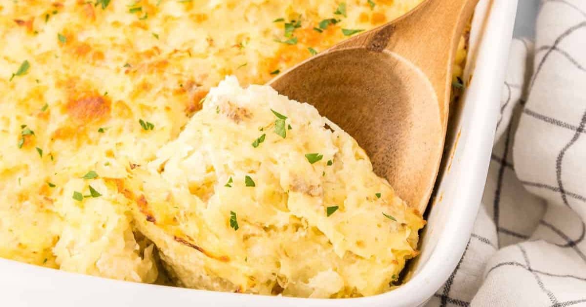 A wooden spoon serving a portion of baked cheesy hash brown casserole garnished with parsley from a white dish on a checkered cloth.