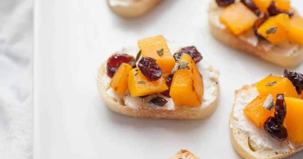 Small slices of bread topped with cream cheese, diced butternut squash, dried cranberries, and herbs on a white plate.