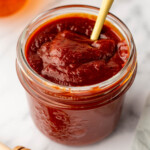 Jar of thick, dark honey barbecue sauce with a spoon, on a marble surface next to a honey dipper and a jar of liquid.