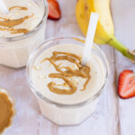 Two glasses of oatmeal smoothie topped with oats and peanut butter drizzle. A banana and sliced strawberry are beside the glasses on a light wooden surface.