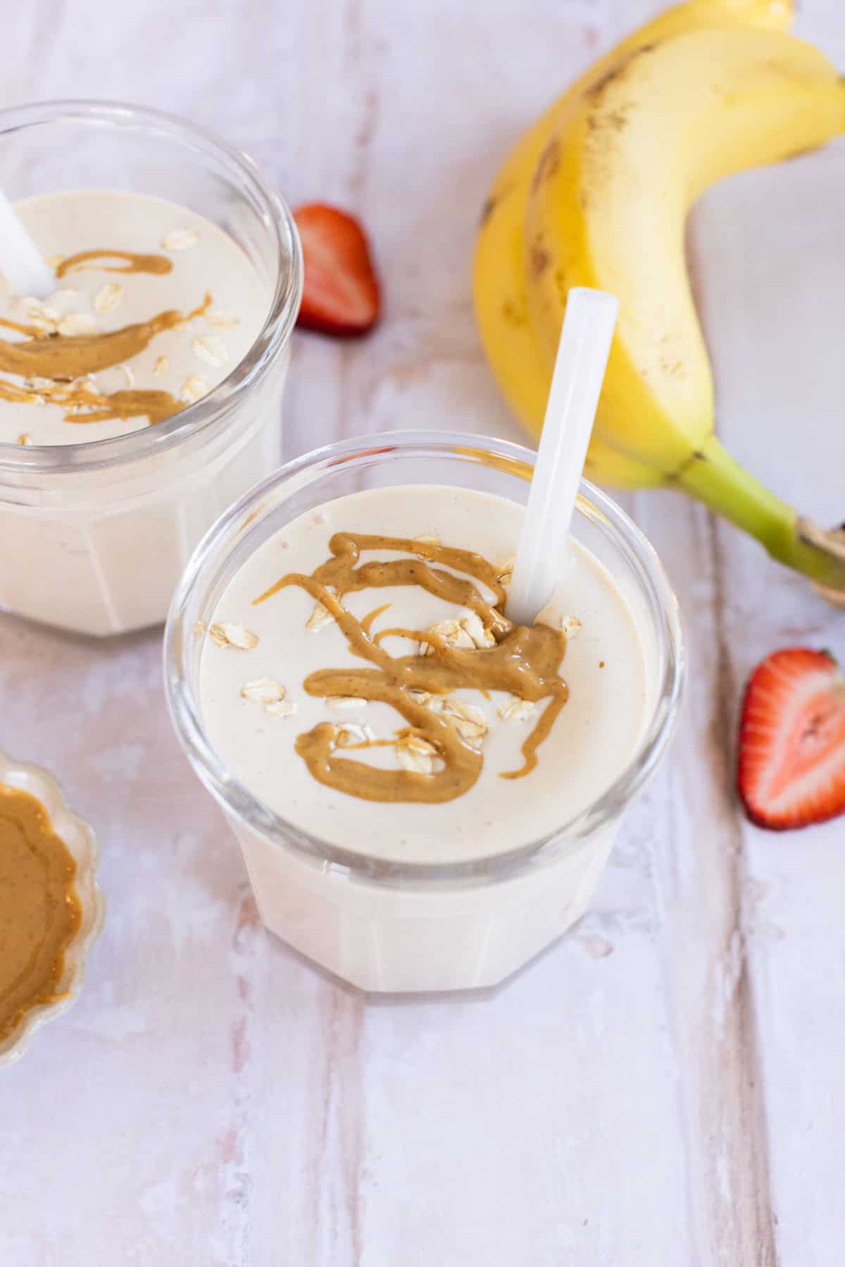 Two glasses of oatmeal smoothie topped with oats and peanut butter drizzle. A banana and sliced strawberry are beside the glasses on a light wooden surface.