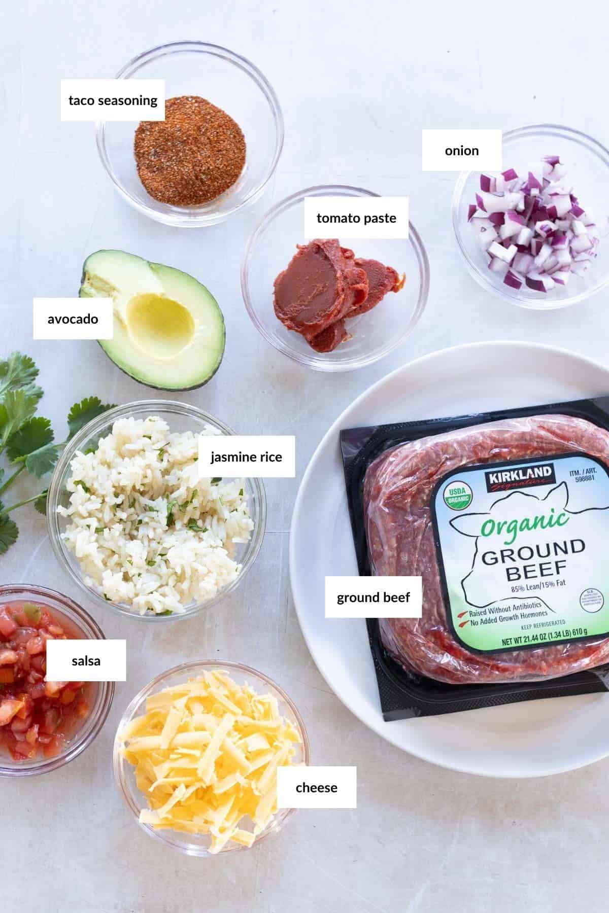 Ingredients including taco seasoning, tomato paste, diced onion, half avocado, jasmine rice, salsa, shredded cheese, and packaged ground beef are arranged on a white surface.