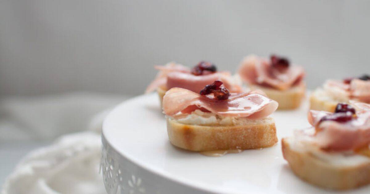 Slices of baguette topped with cheese, prosciutto, and dried cranberries are arranged on a white platter.
