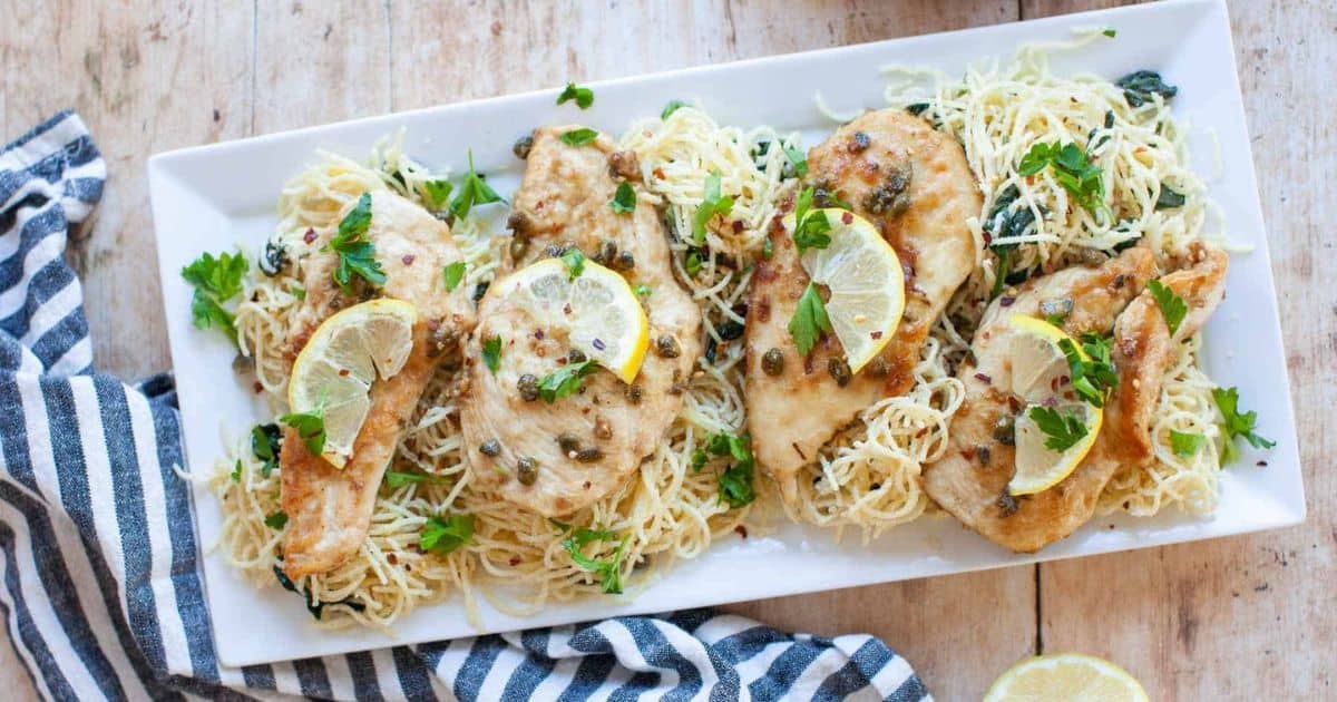 Rectangular white plate with four pieces of chicken breast on a bed of pasta, garnished with lemon slices, capers, and chopped parsley. A striped cloth is partially visible beside the plate.