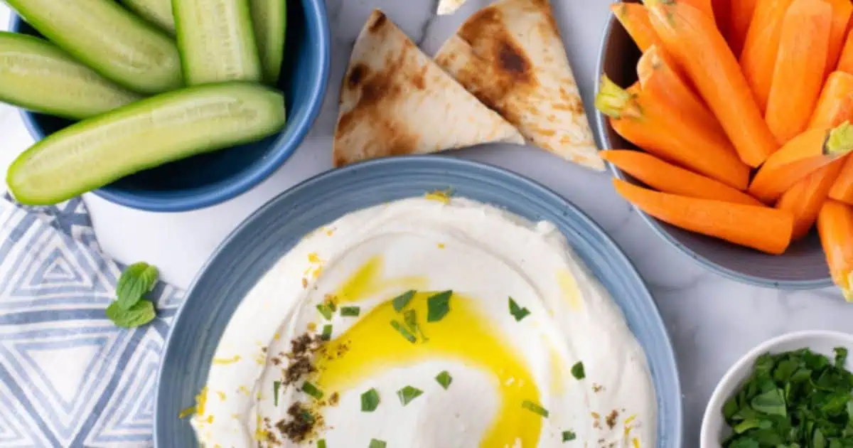 A plate of hummus with olive oil and herbs, surrounded by cucumber slices, carrot sticks, pita bread, and a bowl of parsley on a white table.