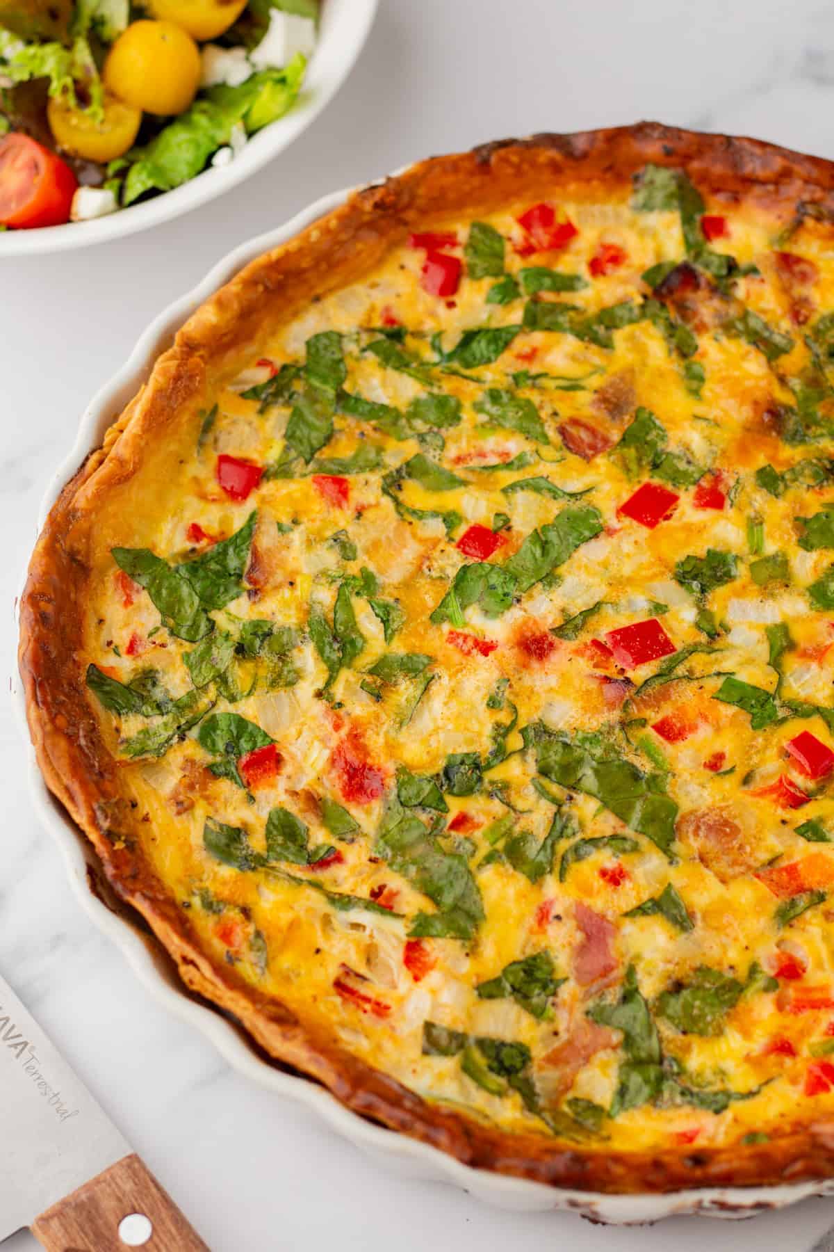 A baked vegetable quiche with visible spinach, red bell peppers, and onions in a pie dish. A bowl of mixed salad is partially visible in the background.