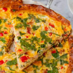 A slice of vegetable quiche with spinach, red peppers, and onions is being served from a pie dish, with a salad in the background.