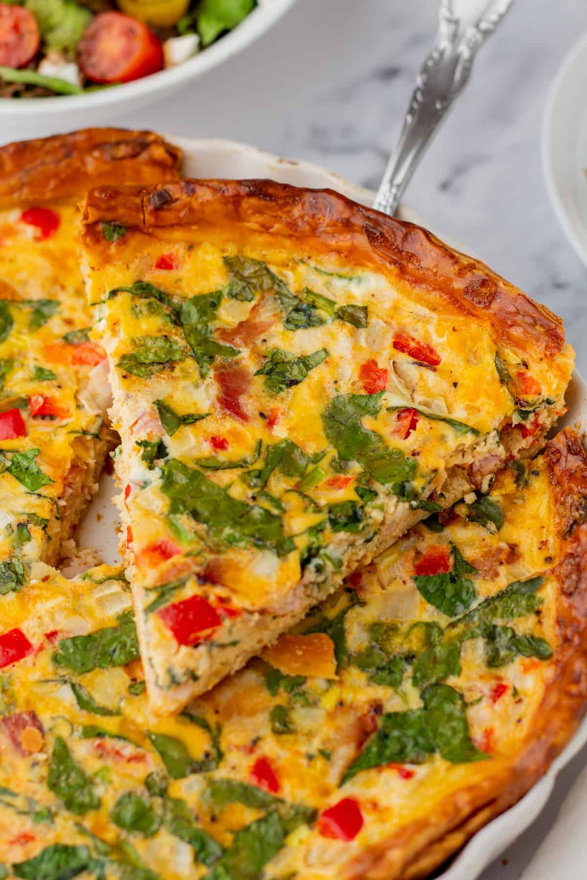 A slice of vegetable quiche with spinach, red peppers, and onions is being served from a pie dish, with a salad in the background.