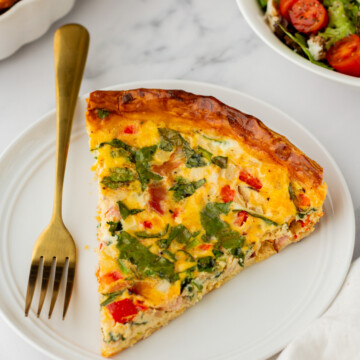 A slice of vegetable quiche on a white plate with a gold fork, next to a bowl of mixed green salad with cherry tomatoes.