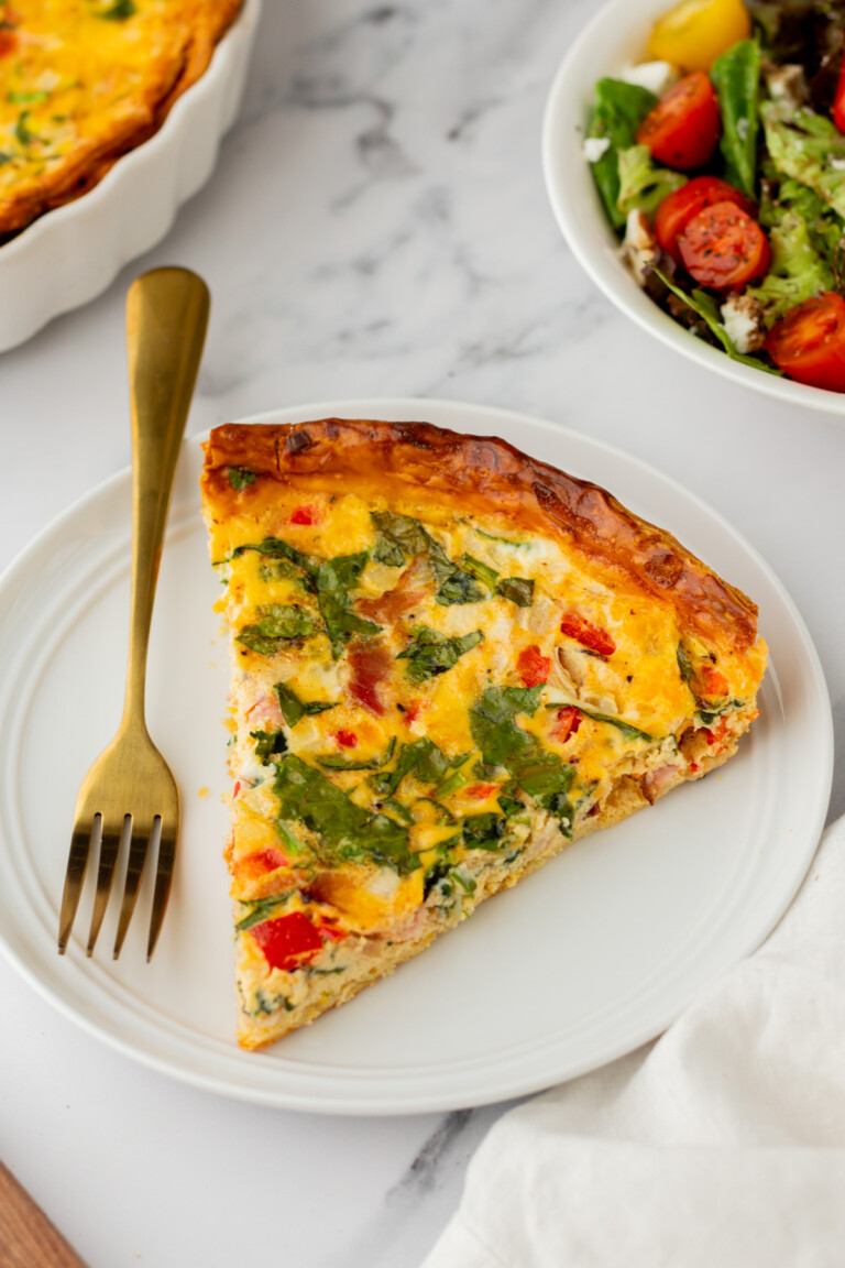 A slice of vegetable quiche on a white plate with a gold fork, next to a bowl of mixed green salad with cherry tomatoes.