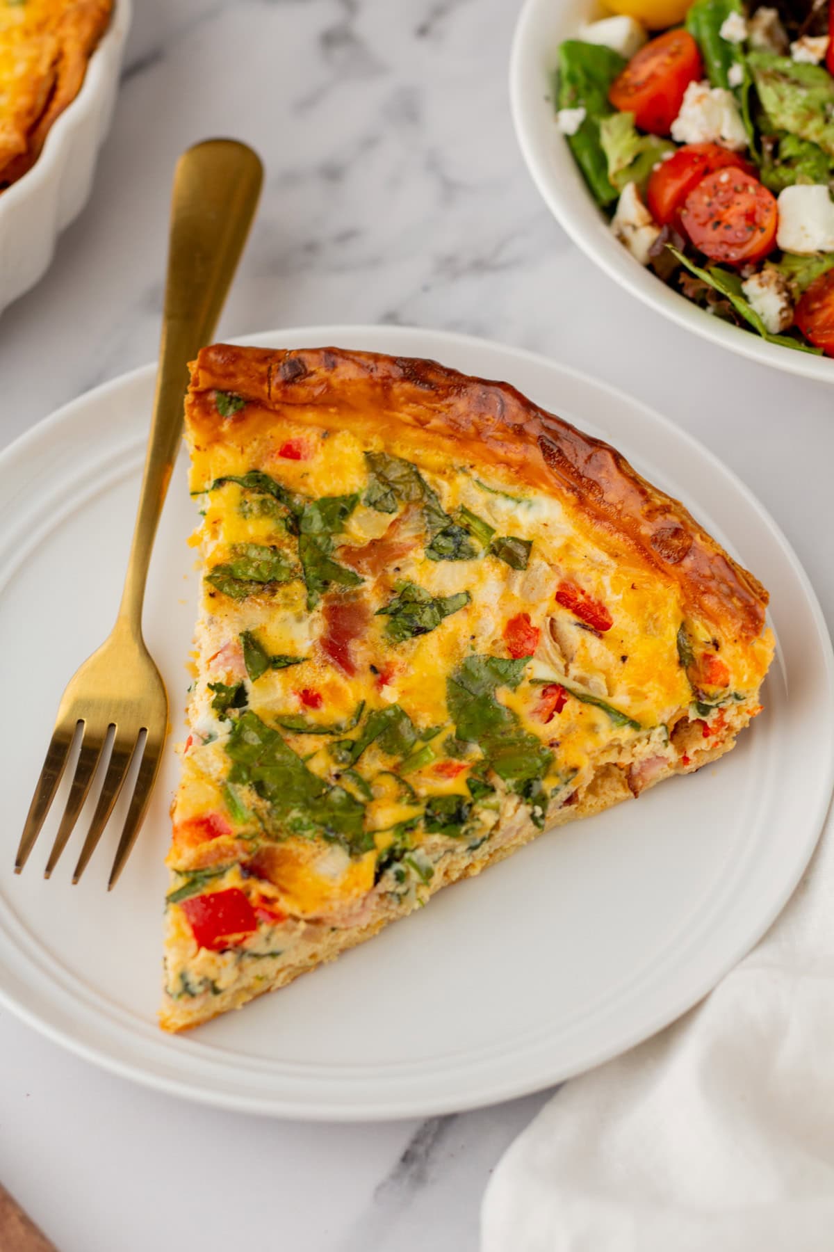A slice of vegetable quiche with spinach, tomatoes, and cheese on a white plate with a gold fork beside a bowl of salad.