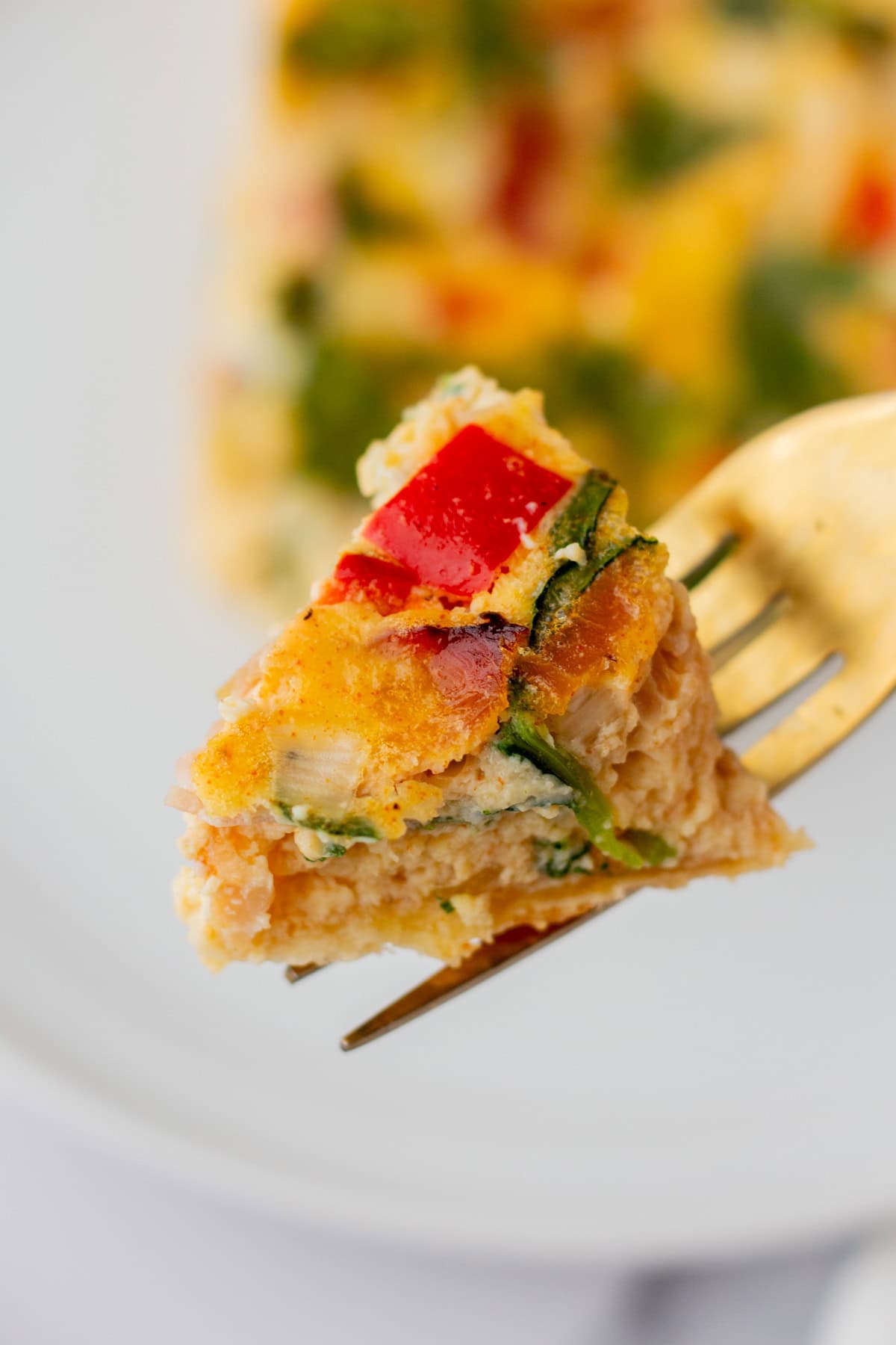 A close-up of a fork holding a bite of baked egg dish with red bell pepper, green vegetables, and a golden-brown crust.