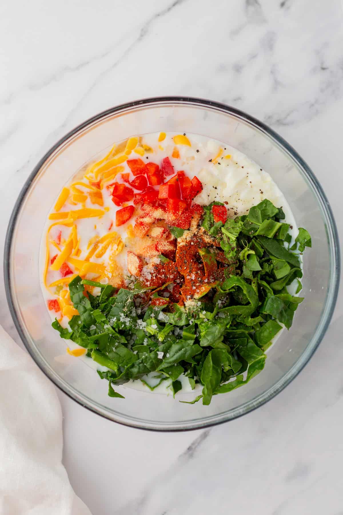 A glass bowl containing shredded cheese, chopped red bell pepper, spinach, yogurt, and spices on a marble surface.