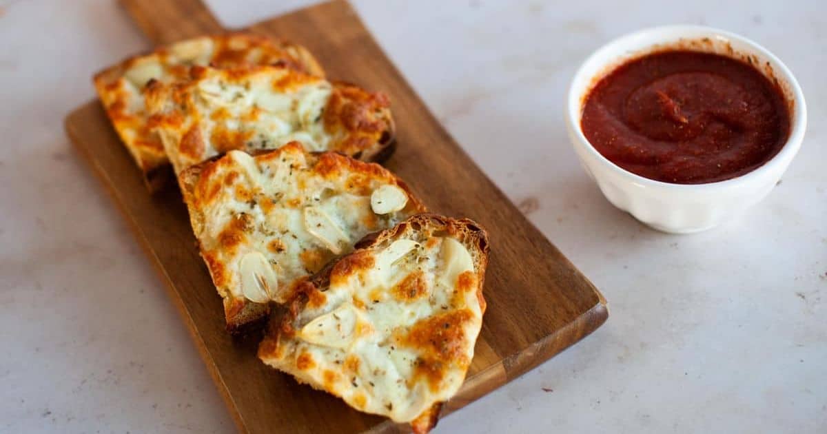 Slices of garlic bread topped with melted cheese on a wooden board, served with marinara sauce in a small white bowl.