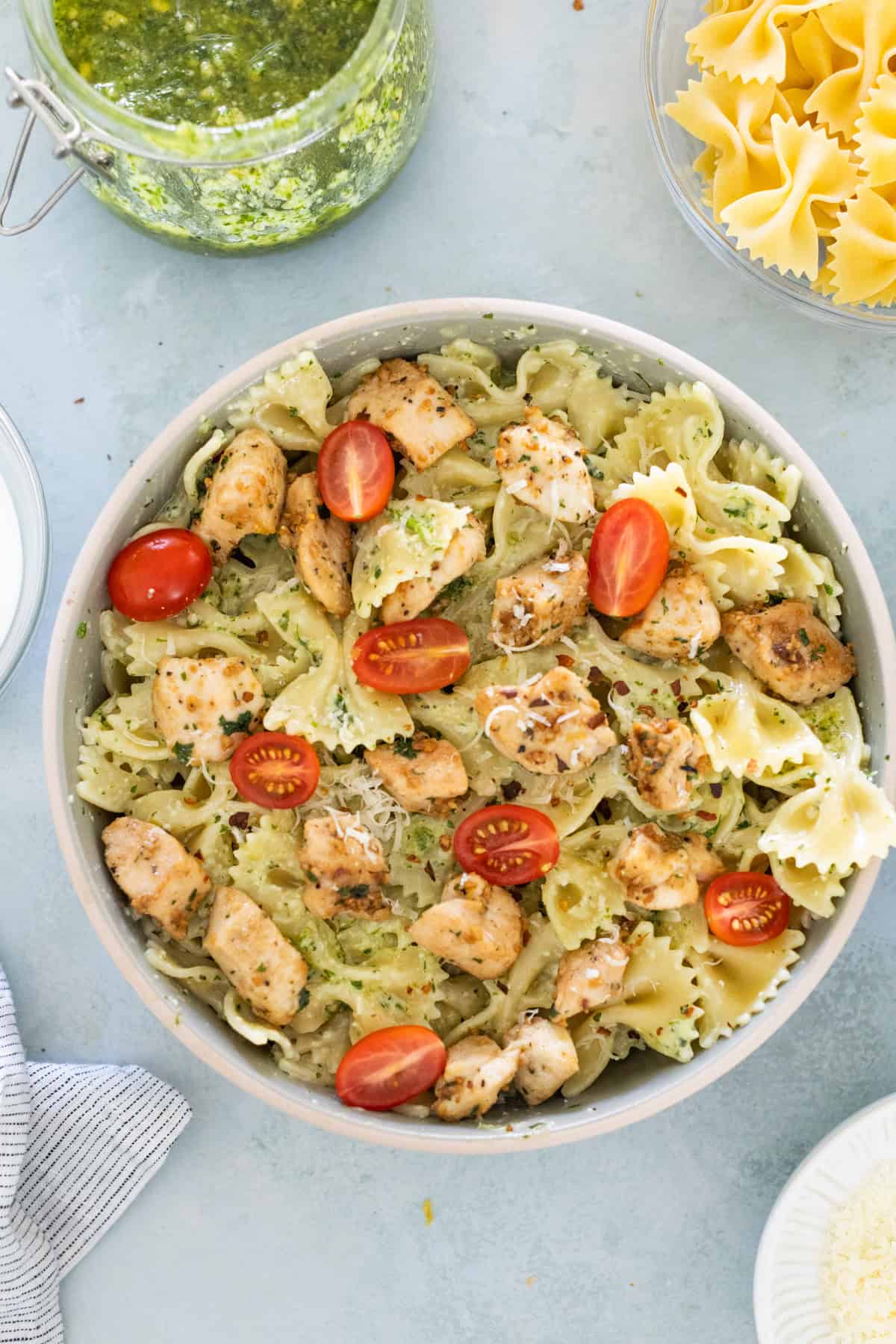 A bowl of bowtie pasta mixed with grilled chicken, cherry tomatoes, and pesto sauce, garnished with Parmesan cheese. A jar of pesto and uncooked pasta are nearby.