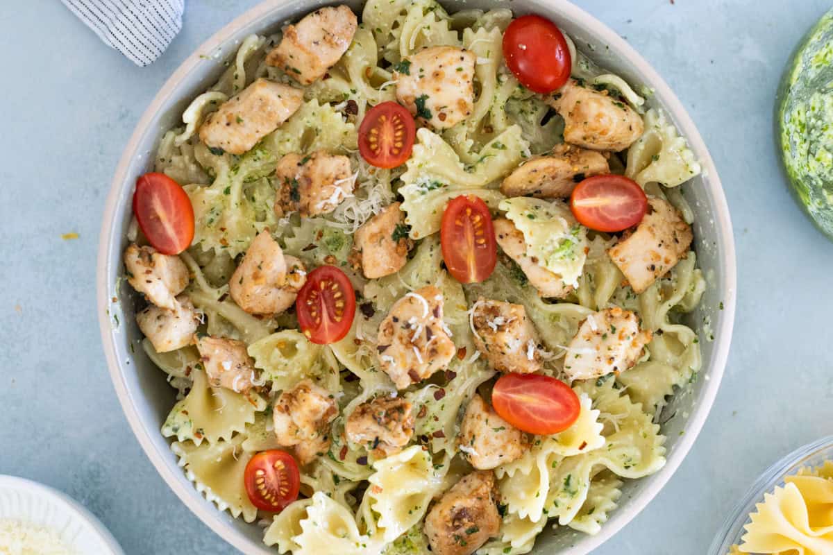 A bowl of farfalle pasta with grilled chicken, cherry tomatoes, and a sprinkle of herbs and cheese, set on a light blue surface.