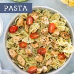A delicious bowl of chicken pesto pasta features bowtie noodles, cherry tomatoes, and grated cheese, artfully surrounded by fresh pesto and uncooked pasta. Text reads "chicken pesto pasta.