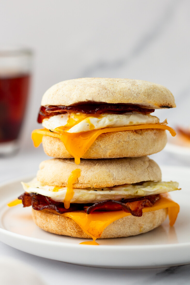 Two breakfast sandwiches stacked on a plate, each with bacon, egg, and melted cheddar cheese between English muffins.