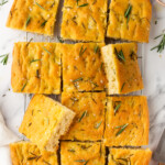 Rectangular focaccia bread cut into twelve pieces, topped with rosemary and coarse salt, resting on a cooling rack with a few herbs scattered around.