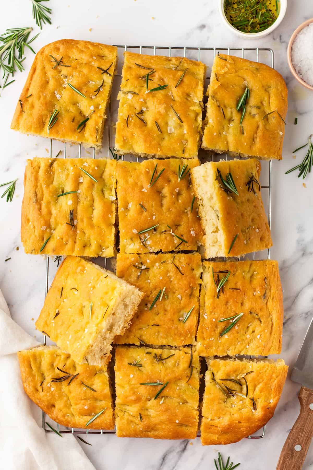 Rectangular focaccia bread cut into twelve pieces, topped with rosemary and coarse salt, resting on a cooling rack with a few herbs scattered around.