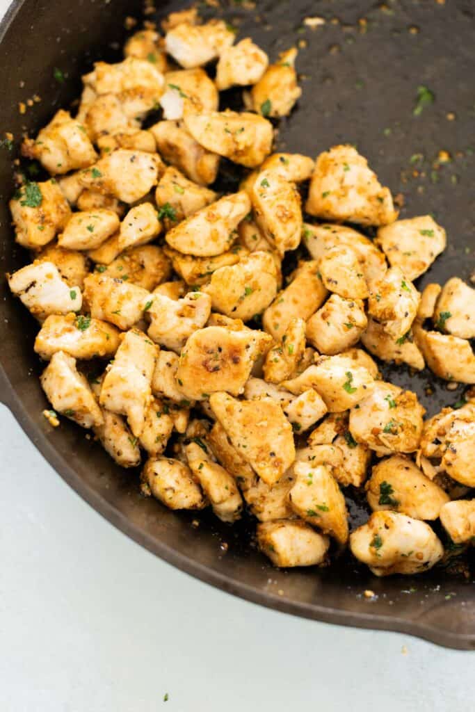 Diced, cooked chicken seasoned with herbs and spices in a black skillet, complemented by a swirl of vibrant pesto.