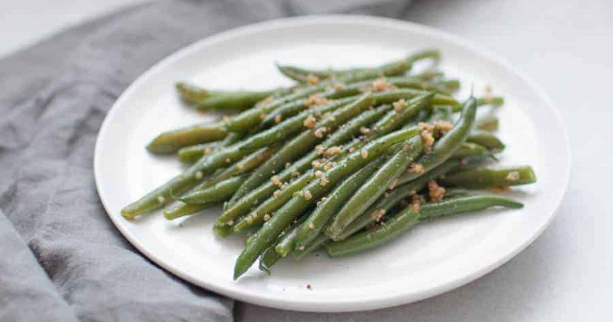 A white plate with cooked green beans topped with minced garlic, placed next to a gray cloth napkin.
