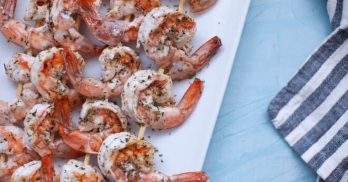 Grilled shrimp skewers seasoned with herbs are arranged on a white rectangular plate next to a striped cloth napkin.