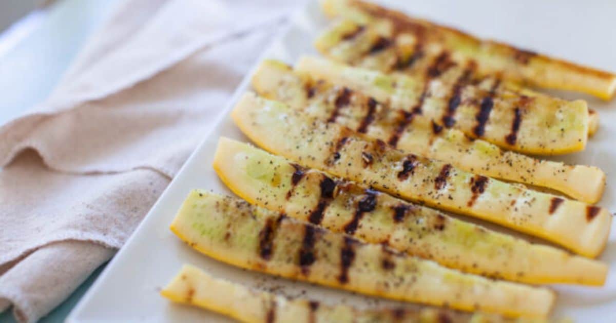 Sliced yellow squash with grill marks arranged on a white rectangular plate next to a folded beige napkin.