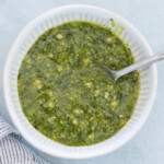 A bowl of green pesto sauce with a spoon inside, placed next to a striped cloth on a light surface.