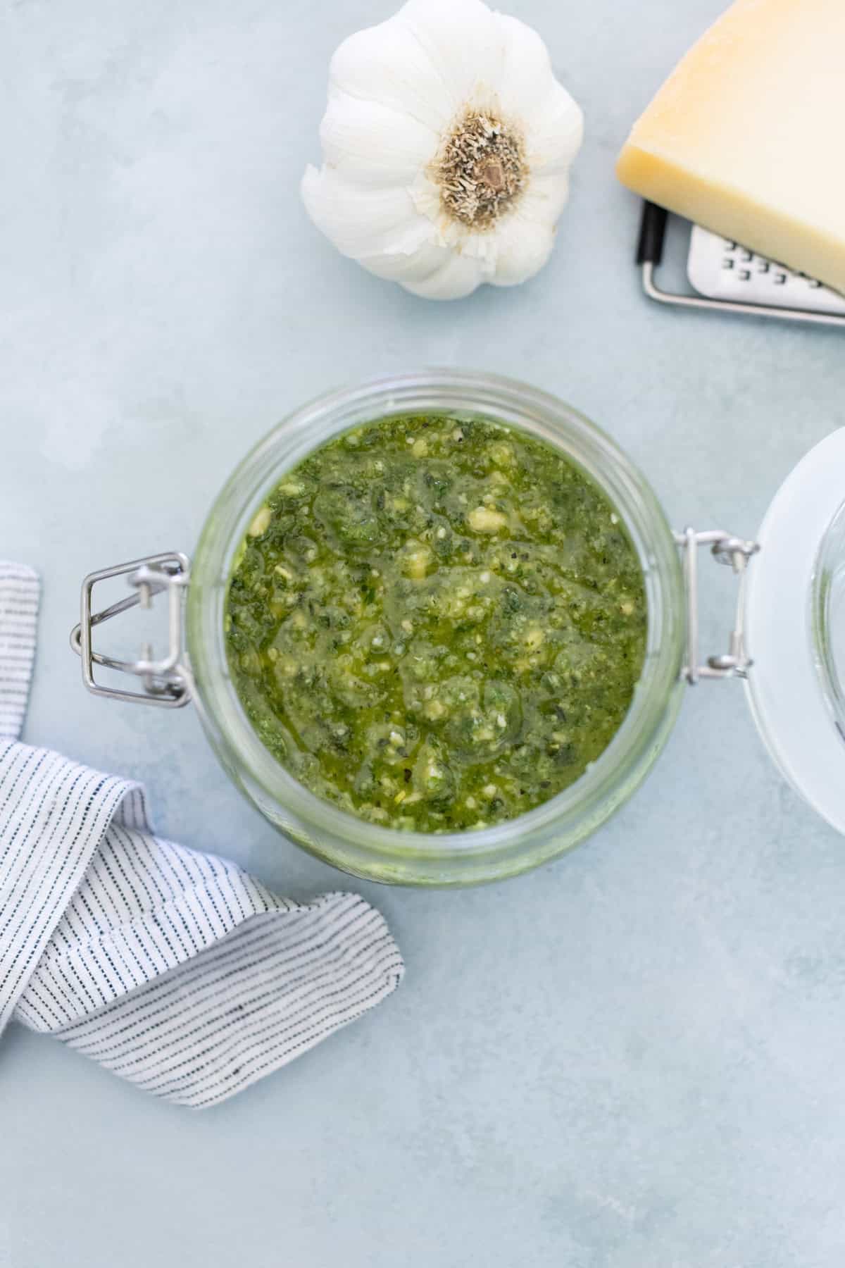 Jar of green pesto sauce on a light surface, surrounded by a garlic bulb, cheese wedge on a grater, and blue-striped cloth.