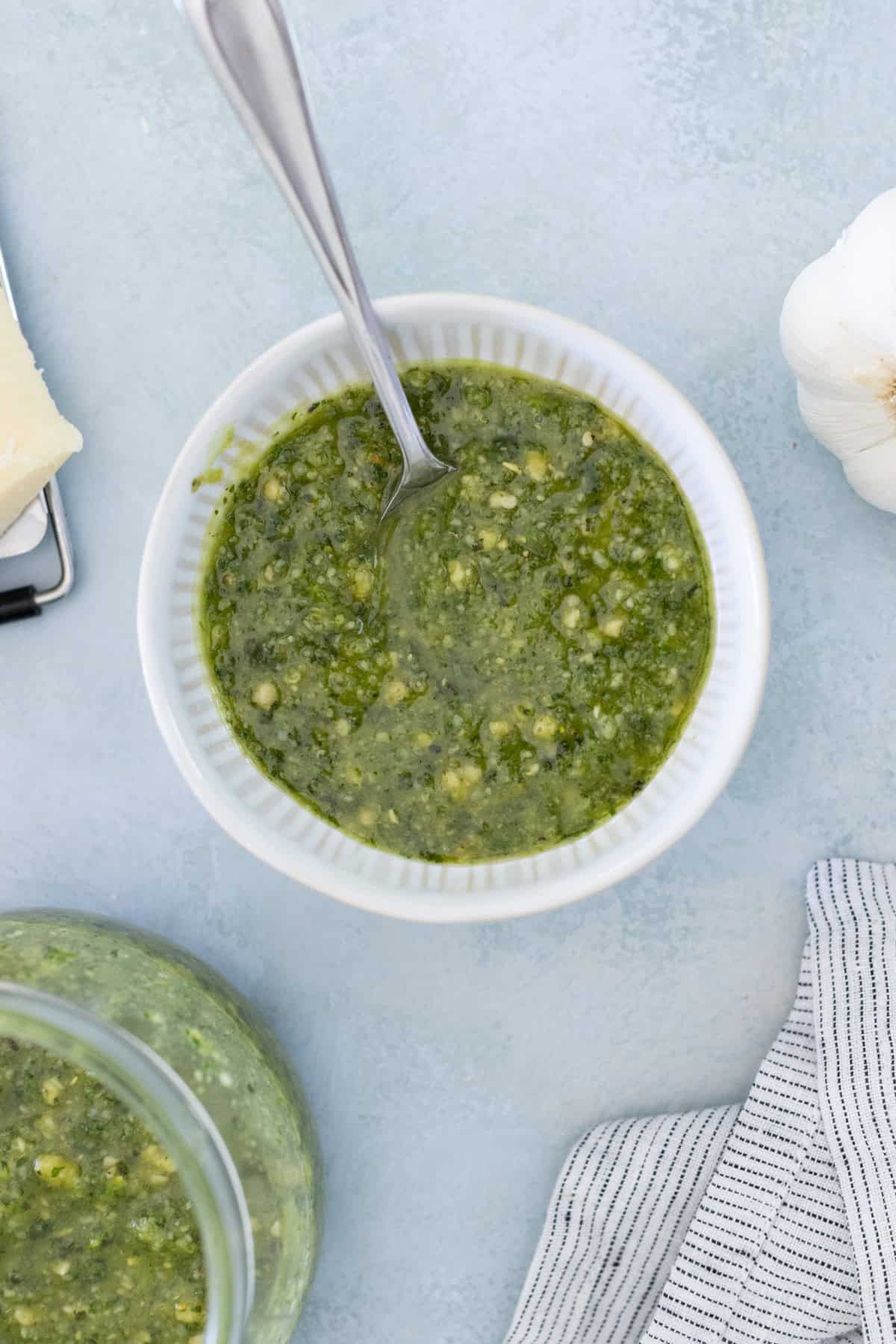 A bowl of green pesto with a spoon inside, placed on a light blue surface. Nearby is a garlic bulb, a piece of cheese, and a striped cloth.