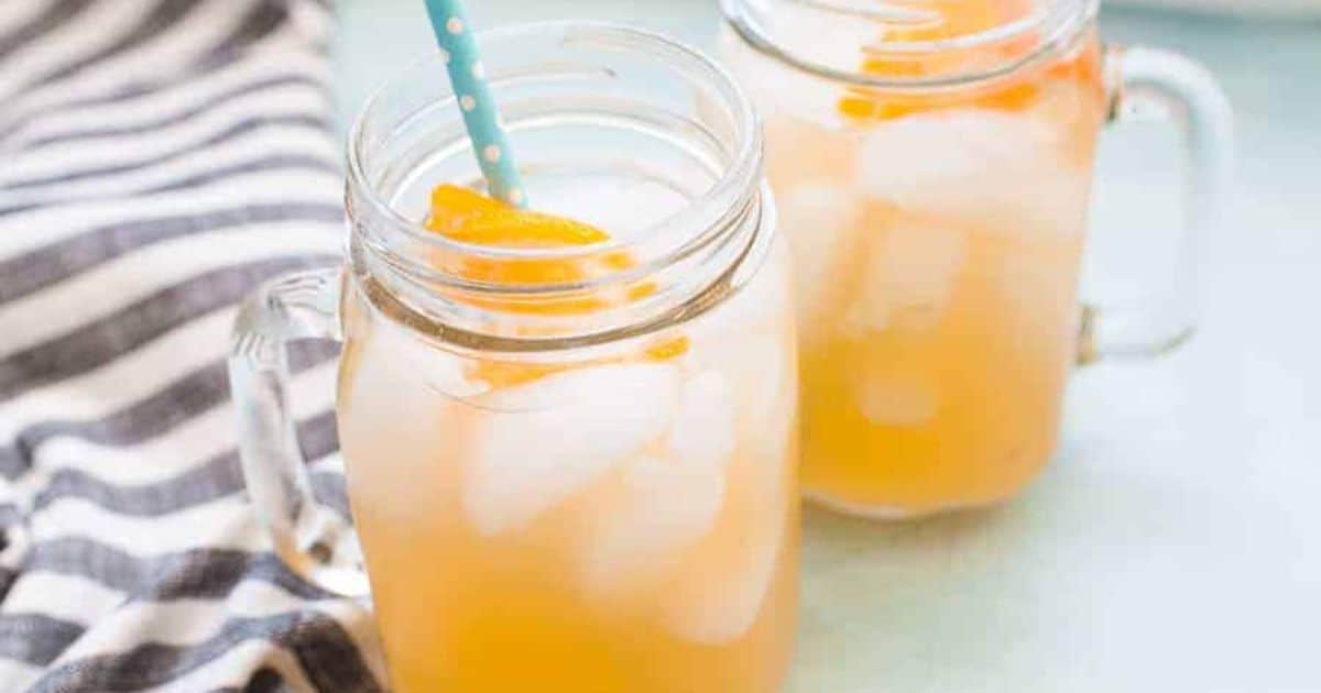 Two mason jar glasses filled with iced orange-colored beverage and orange slices, one with a blue polka dot straw, next to a striped cloth.