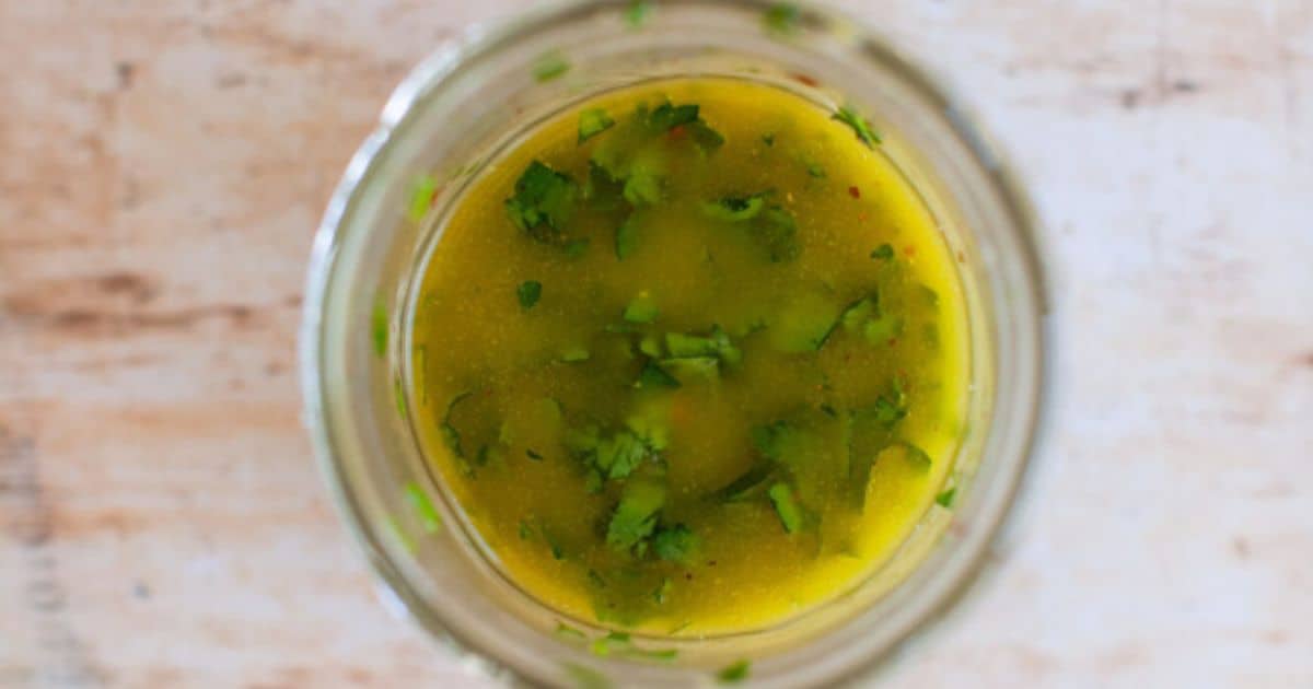 A glass jar filled with yellow-green salad dressing, garnished with chopped fresh herbs, viewed from above on a light surface.