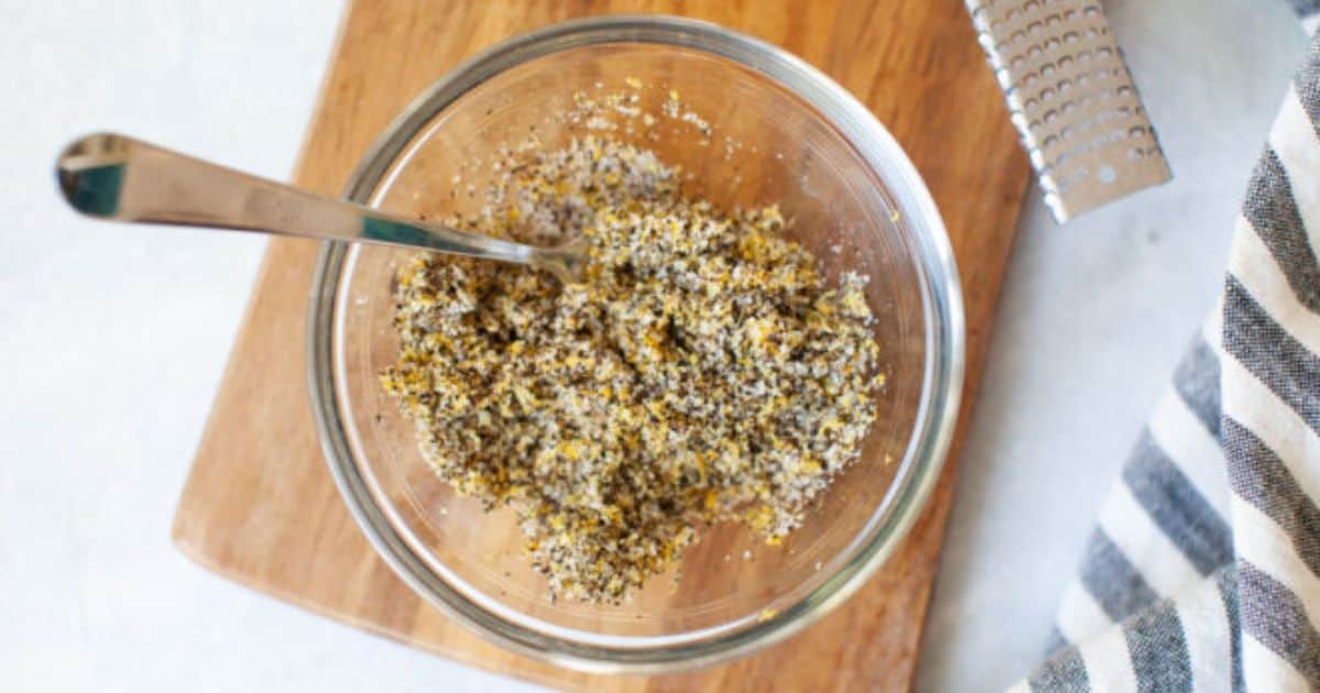 A glass bowl filled with a coarse mixture of black pepper and lemon zest, with a metal spoon, on a wooden cutting board next to a striped cloth and a zester.
