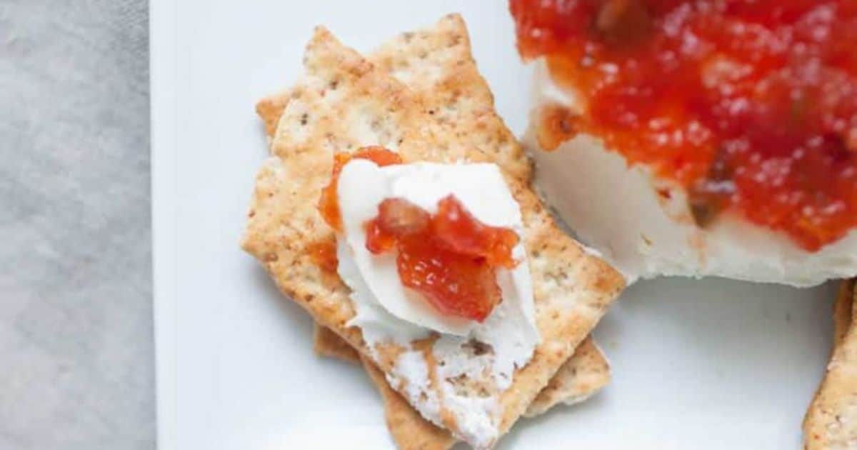 Two crackers topped with cream cheese and red salsa are placed on a white surface next to a larger portion of cream cheese covered with salsa.