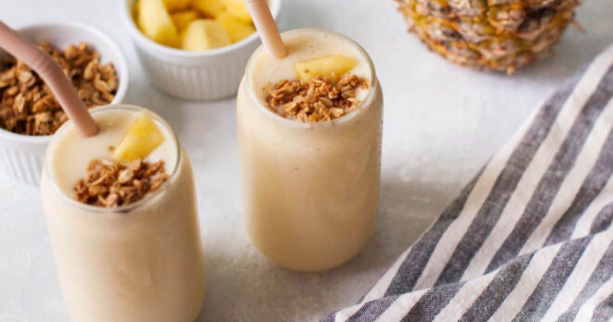 Two glasses of pineapple smoothie topped with granola and pineapple chunks, with straws, sit on a table near a striped cloth and small bowls of granola and pineapple.