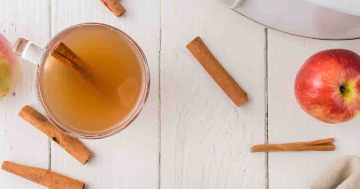 A cup of apple cider with a cinnamon stick, whole cinnamon sticks, and a red apple on a white wooden surface.
