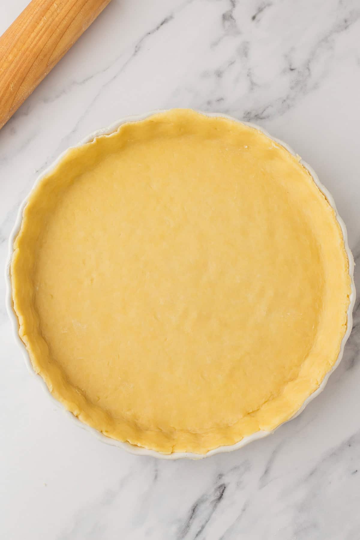 A round tart pan filled with an unbaked pastry crust sits on a marble surface next to a wooden rolling pin.